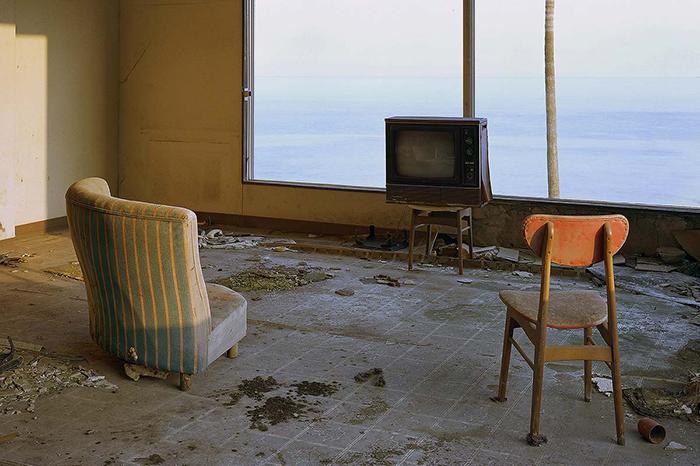 Two empty chairs face an old TV box. The window behind the TV shows the sea. Photo by Max Ernst Stockburger.