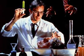 Jeffrey Combs in a publicity still for RE-ANIMATOR, playing the mad scientist, Herbert West, wielding a glowing syringe over a decapitated head.
