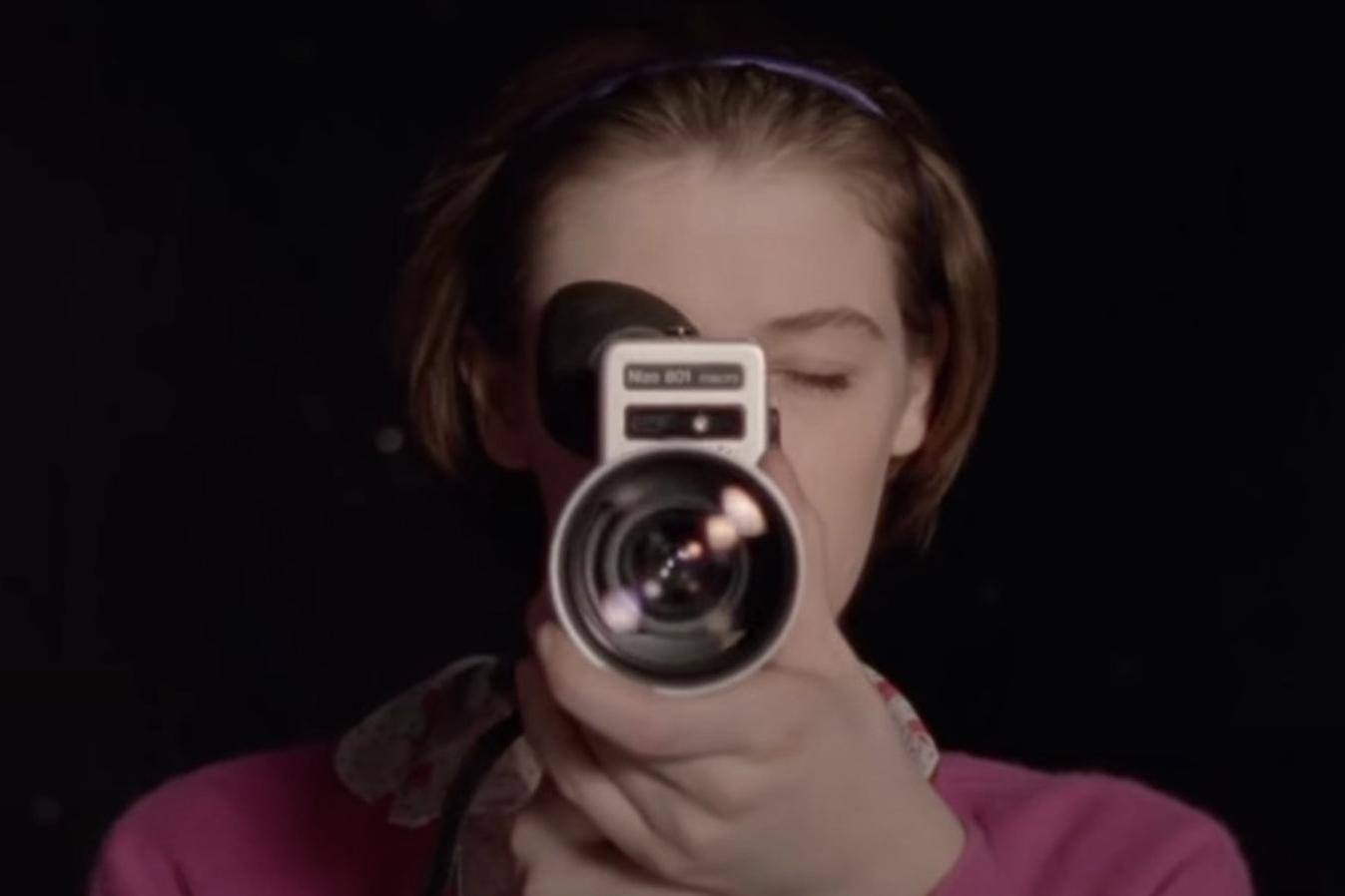 Film still from THE SOUVENIR PART II, showing Honor Swinton Byrne as Julie, looking through a hand-held movie camera at us.