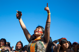 Indigenous woman raises hands aloft while holding a cell phone, surrounded by other women.