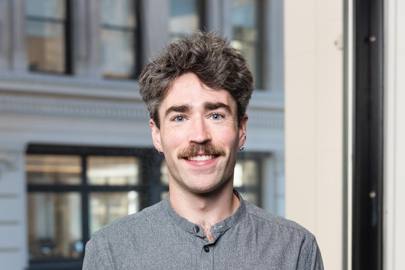 Researcher stands in a gray soft collar shirt with blue eyes in front of a building