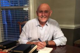 John Evans sits at a desk with a stack of journals. He has a pen in his hand as he begins to write. He smiles at the camera. John Evans sits at a desk with a stack of journals. He has a pen in his hand as he begins to write. He smiles at the camera.