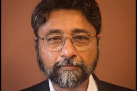 Anjan looks directly at the camera wearing a black suit and white button down. He wears glasses and has a full beard and mustache.