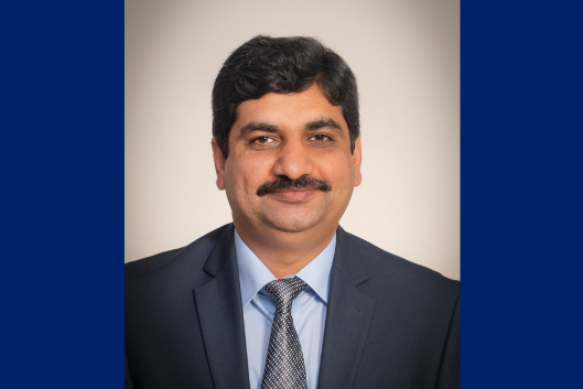 Formal headshot of seminar speaker, Dr. Salik Hussain, wearing a navy blue suit and tie in front of a light gray background