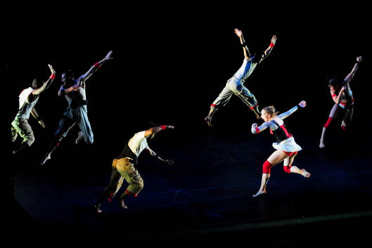 6 dancers in various stages of movement on a darkened stage.