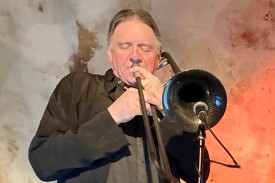 Conrad Herwig playing the trombone against a gray and orange wall.