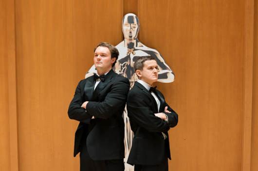 Two young men in formal wear standing in front a a human size Oscar statue