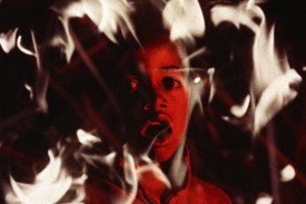 Still image of close-up on a boy's face, tinted red and surrounded by flames from Time of the Heathen