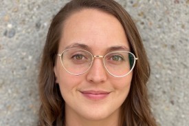 Researcher stands in a black and white polka dot shirt in glasses against a concrete wall