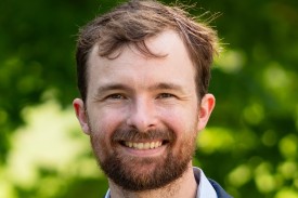 Researcher stands in a navy blue jacket and light blue shirt in an outdoor setting