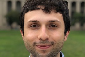 A researcher with dark hair and short beard stands in an outdoor setting wearing a light blue pin stripe shirt