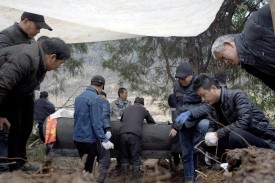 People gathered around an exhumed casket under a protective tarp. Background view of the village countryside, seen through the branches of trees.