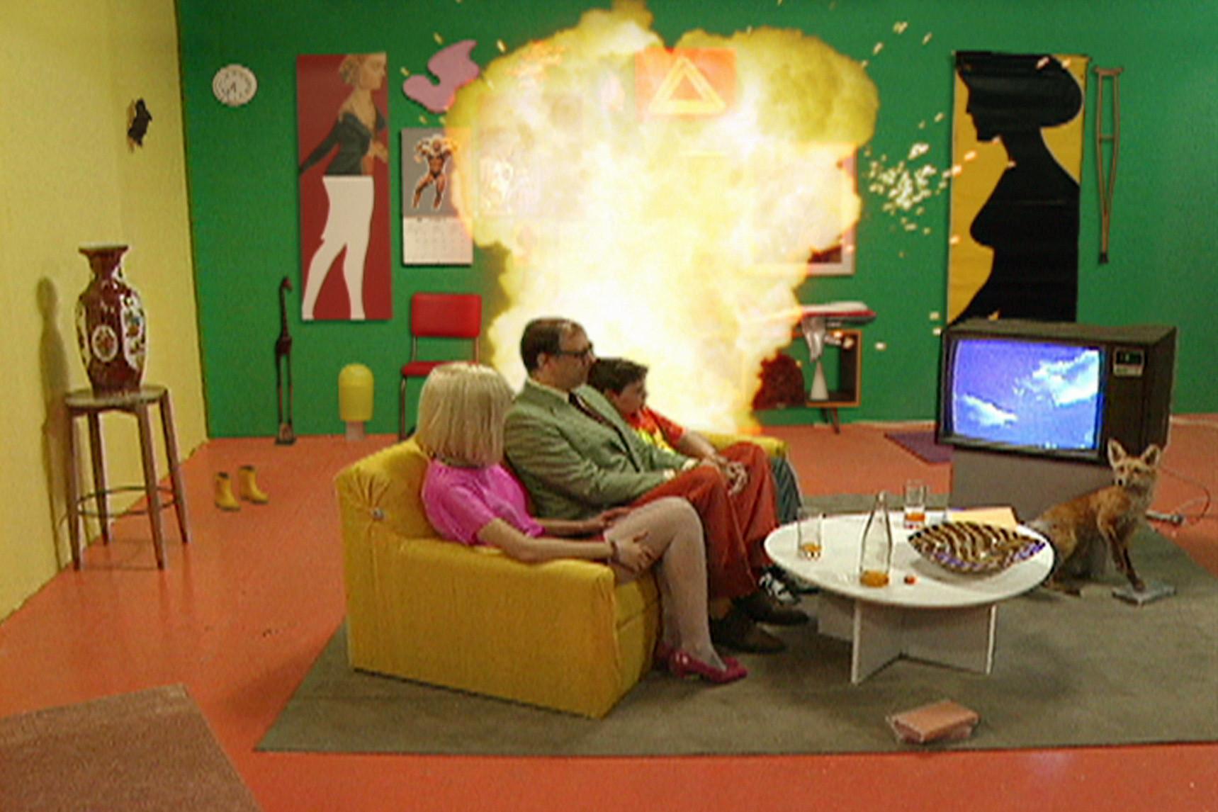A sitcom-style family sits on a couch calmly gazing at a TV while an explosion goes off behind them, in a still from *CORPUS CALLOSUM A sitcom-style family sits on a couch calmly gazing at a TV while an explosion goes off behind them, in a still from *CORPUS CALLOSUM