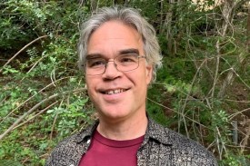 Eric Schwitzgebel - Man, with glasses, wearing maroon t-shirt and black and white collard shirt standing in front of greenery