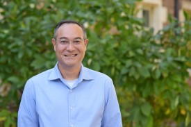 A man smiles, standing in front of a wall with leaves.