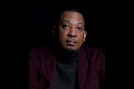 A man wearing a black turtleneck and deep-purple sportcoat poses against a black background.