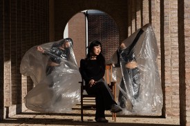 A woman wearing black clothes sits on a chair with a Duke arch behind her and clones of herself in plastic beside her A woman wearing black clothes sits on a chair with a Duke arch behind her and clones of herself in plastic beside her