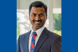 Dr. Satya Achanta, smiling and wearing a dark blazer and red and white striped tie.